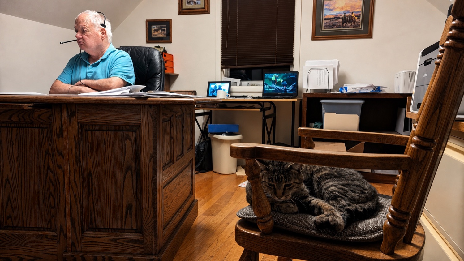 Mike at his desk in the upstairs office wearing a phone headset, papers in front of him, while Goonhead sleeps curled on a wooden rocking chair beside the desk.