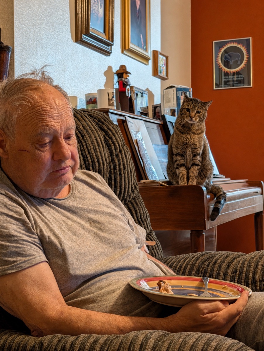 Mike holding a dinner plate on his lap, looking sideways at Goonhead, who sits upright on the piano staring back at him.