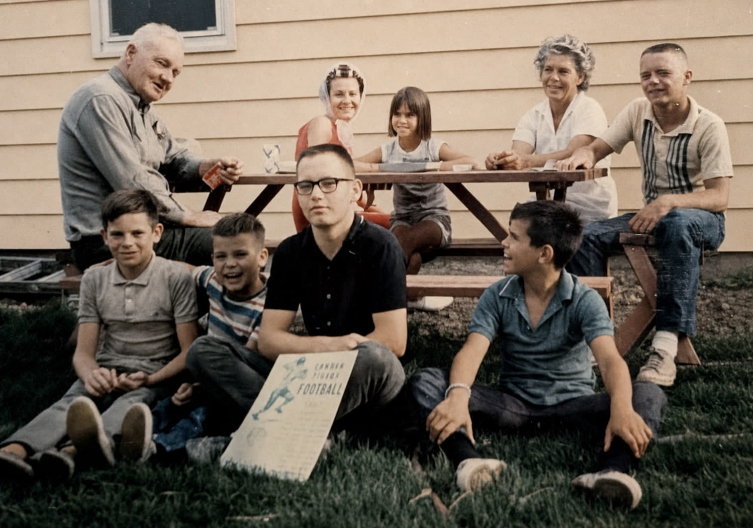 Nine members of the Anderson family and their relatives outside the newly finished house in 1967 &mdash; four adults seated at a picnic table against the siding, and four boys and a young girl on the grass in front, one of the boys holding a football program.
