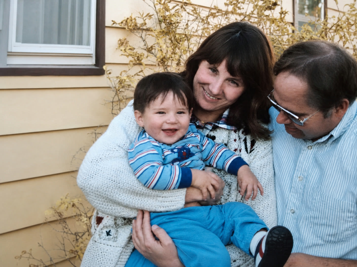 Saundra holding baby Matt on her lap outside the house, both smiling, with Mike leaning in beside them looking down at his son.