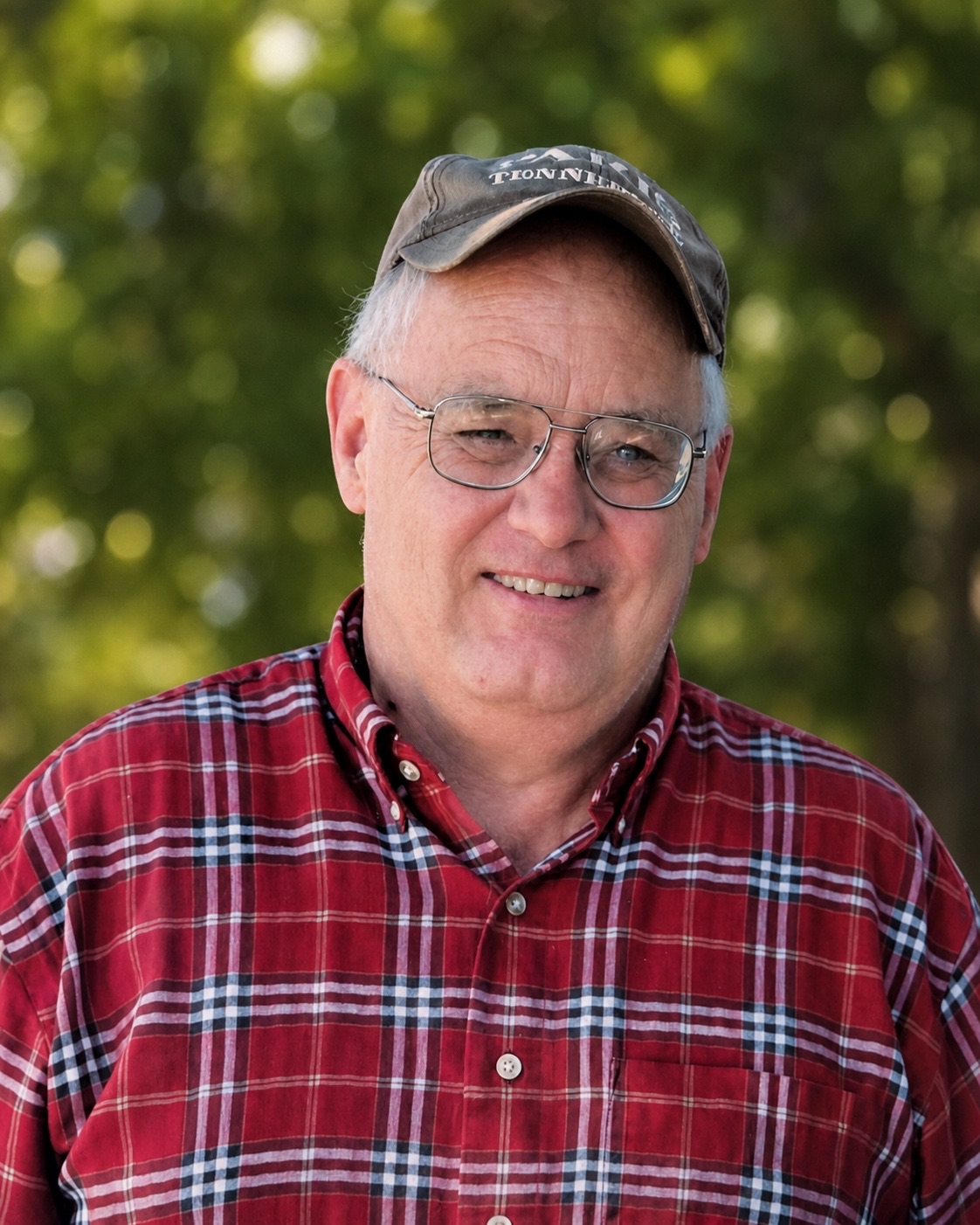 Mike Anderson smiling in a red plaid shirt and a well-worn ball cap, green trees behind him.