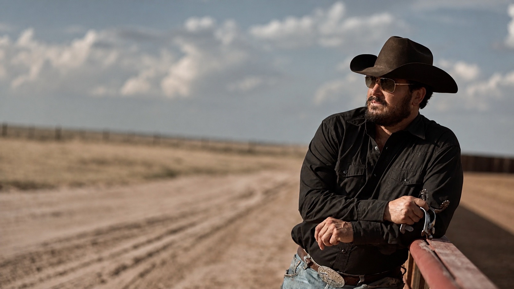 A cowboy in a black hat leaning on a fence rail, looking out over a dirt road and open plain.